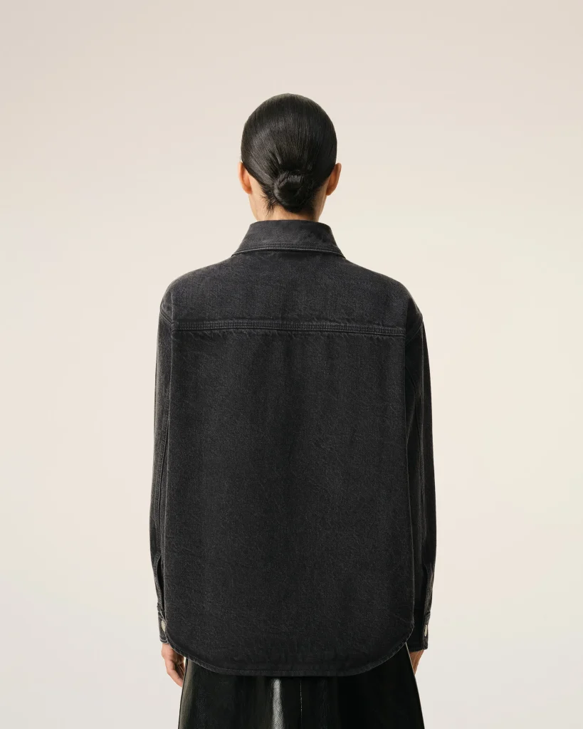 Back view of a person with dark hair in a neat bun, wearing a black denim jacket against a pale background.