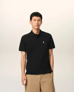 Person standing against a light beige background, wearing a black polo shirt and tan khaki pants with a neutral expression.