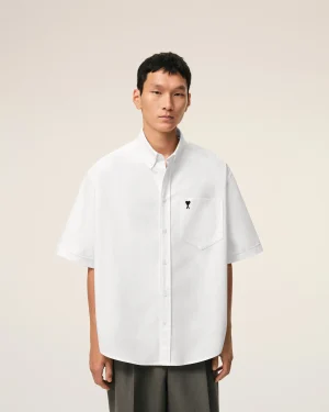 Young man wearing an oversized white button-down shirt, standing against a light beige backdrop.