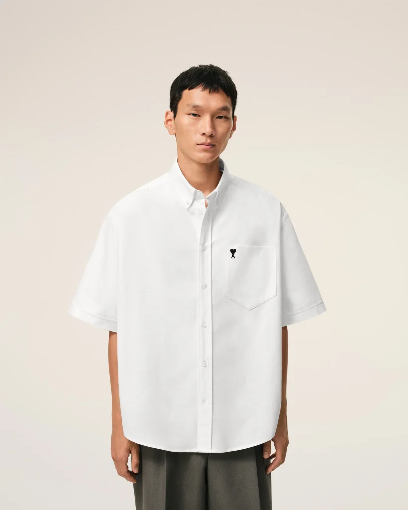 Young man wearing an oversized white button-down shirt, standing against a light beige backdrop.