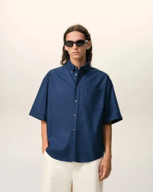 Person wearing a dark blue button-down shirt and white pants, sunglasses, standing against a pale beige background.