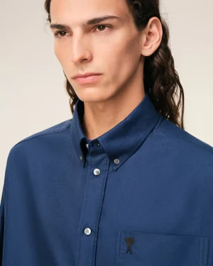 Close-up of a person wearing a blue button-down shirt with a small heart-shaped emblem on the left chest pocket, face partially visible.