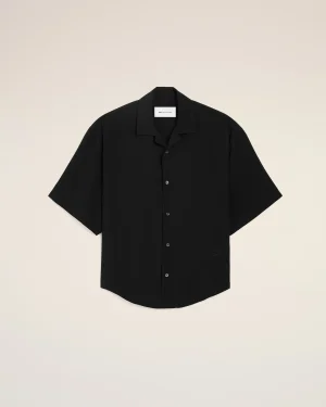 Flat lay of a black short-sleeve button-down shirt on a beige background, tag visible at the collar