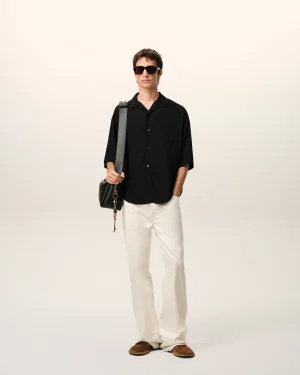 A man in a black short-sleeve shirt, white pants, brown loafers, and dark sunglasses stands with a gray shoulder bag against a light background.