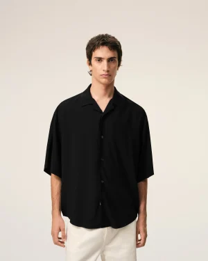 Young man with short dark hair wearing a black button-up shirt, standing against a light beige background.