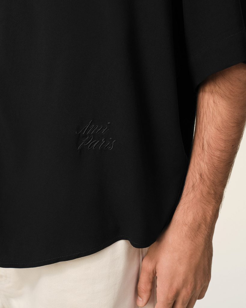 Close-up of a black garment with subtle cursive embroidery near the hem and a forearm resting at the side with beige pants visible.