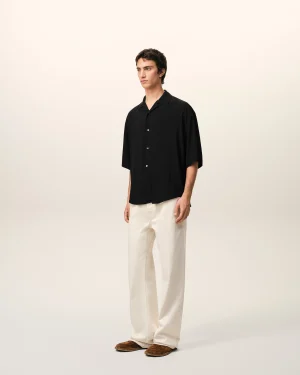 Person wearing a black button-up shirt, cream pants, and brown loafers stands against a light beige studio backdrop.