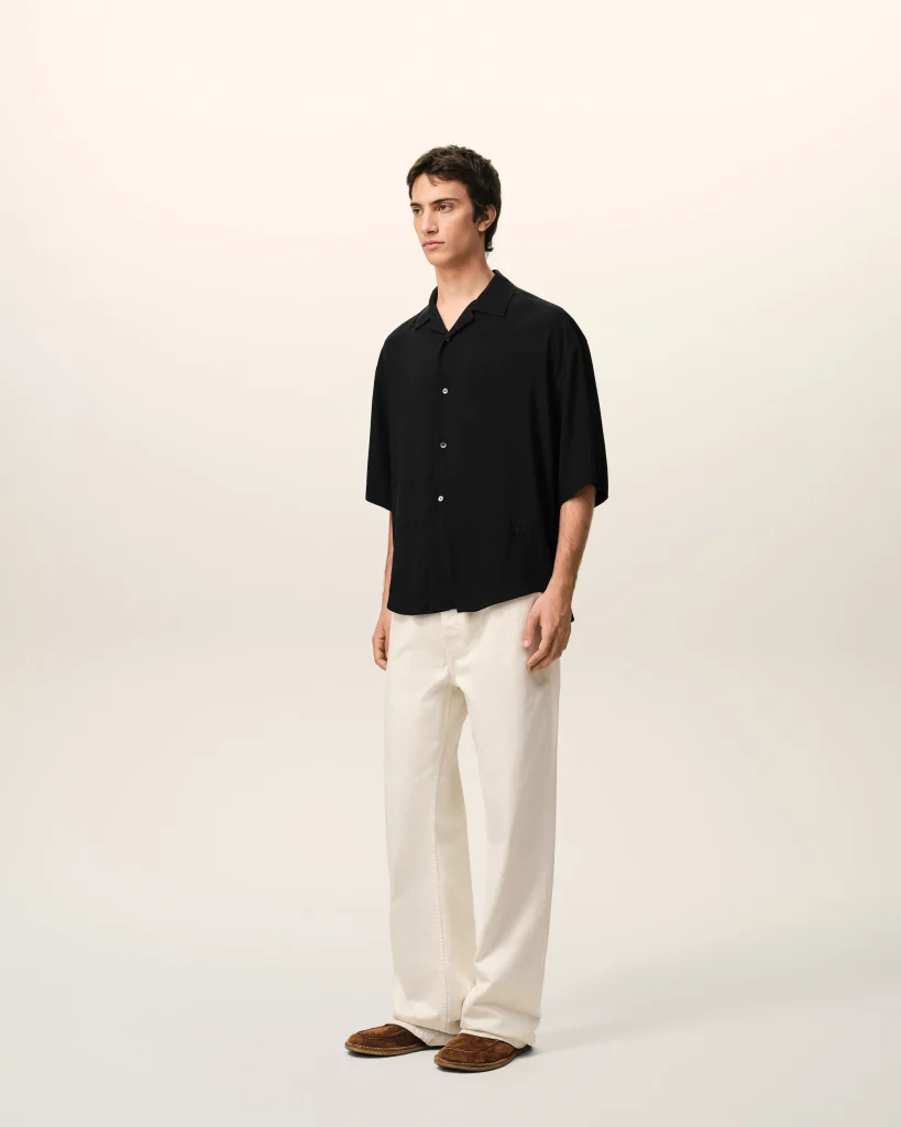 Person wearing a black button-up shirt, cream pants, and brown loafers stands against a light beige studio backdrop.