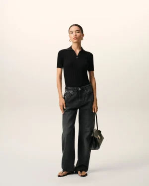 Model standing against a light beige background, wearing a black ribbed polo top, dark jeans, gold hoop earrings, and carrying a small black handbag with a strap.