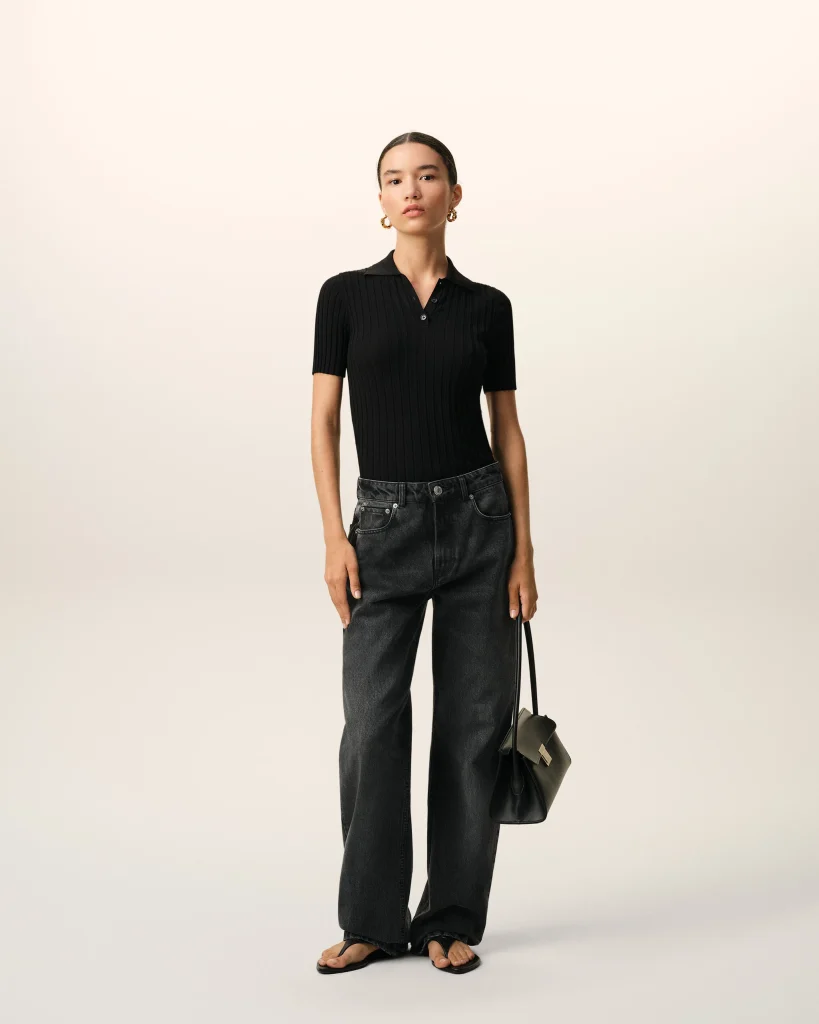 Model standing against a light beige background, wearing a black ribbed polo top, dark jeans, gold hoop earrings, and carrying a small black handbag with a strap.