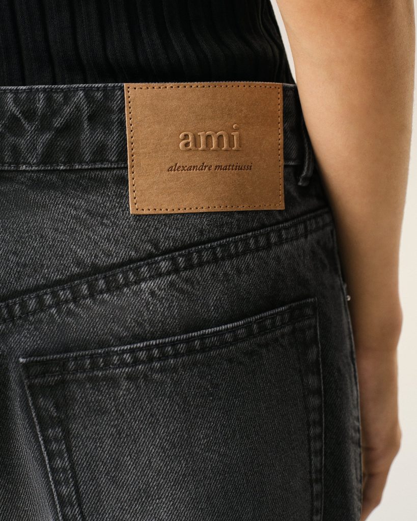 Back of dark denim jeans with a tan leather brand patch reading "ami" and "alexandre mattiussi" near the waist.