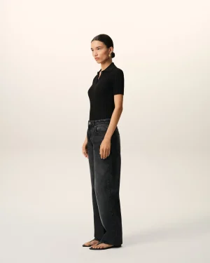 Profile view of a woman standing in a black ribbed polo shirt and dark jeans, wearing gold earrings, against a light neutral backdrop.