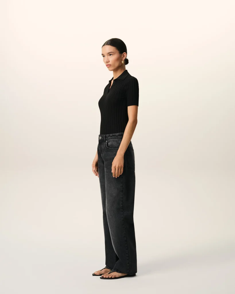 Profile view of a woman standing in a black ribbed polo shirt and dark jeans, wearing gold earrings, against a light neutral backdrop.