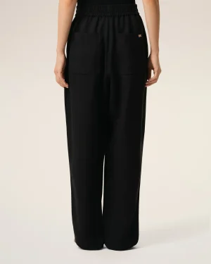 Back view of a person wearing black wide-leg pants with a gathered elastic waistband and large front pockets, small metal tag on the right pocket edge.