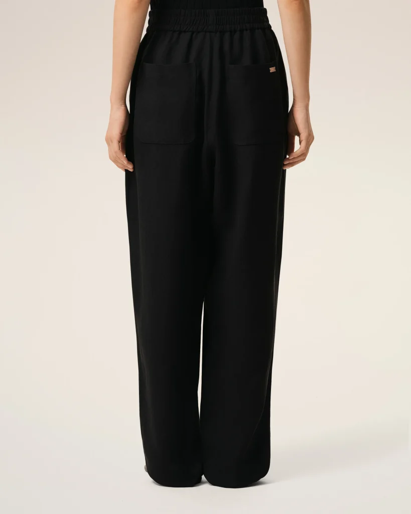 Back view of a person wearing black wide-leg pants with a gathered elastic waistband and large front pockets, small metal tag on the right pocket edge.