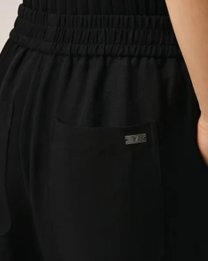 Close-up of black fabric pants with a wide, elastic waistband and a small silver metal tag featuring a heart emblem near the pocket.