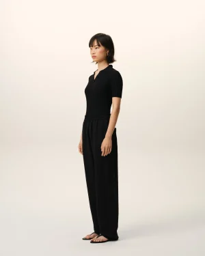 Person standing in profile wearing a black ribbed polo shirt and wide-leg black pants against a pale gradient backdrop.