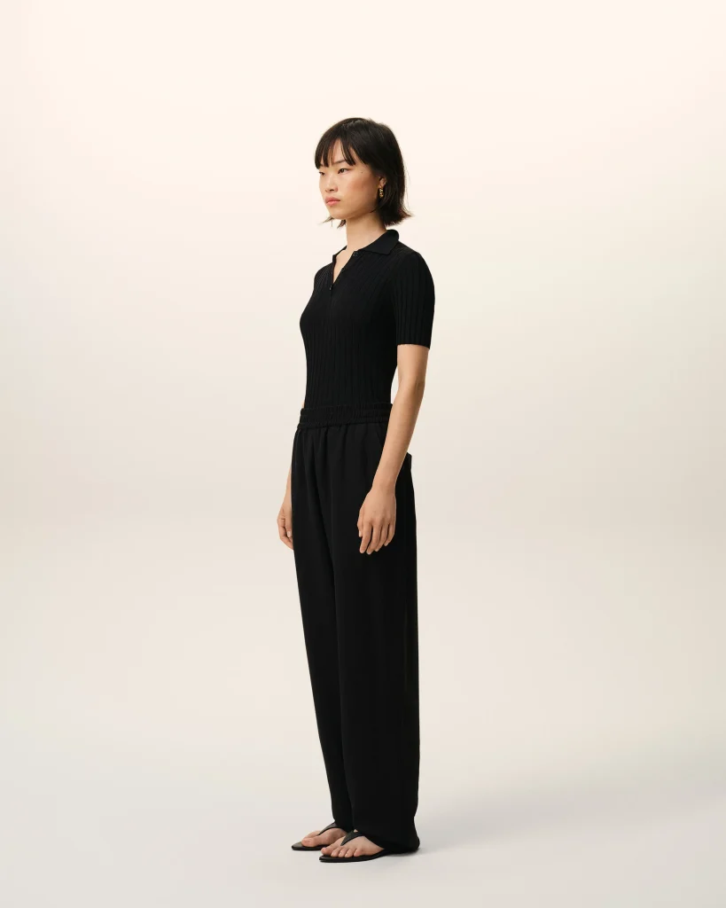 Person standing in profile wearing a black ribbed polo shirt and wide-leg black pants against a pale gradient backdrop.