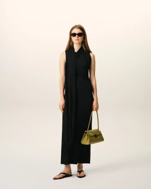 Full-body shot of a woman in a black sleeveless dress and sunglasses, holding an olive-green handbag, standing against a pale gradient background.