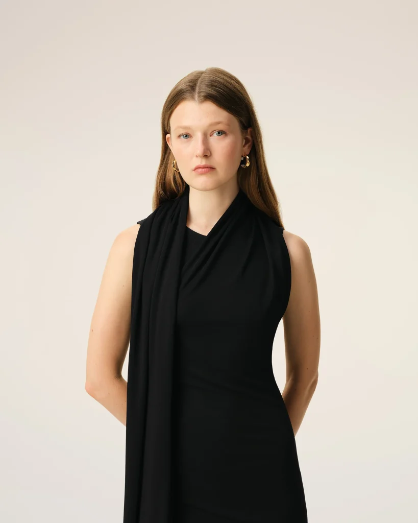 Portrait of a woman with long brown hair wearing a black draped sleeveless top, looking directly at the camera against a pale beige studio backdrop.