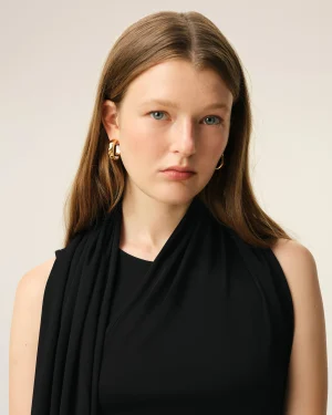 Close-up portrait of a woman with blue eyes, wearing gold hoop earrings and a black draped top against a beige background.