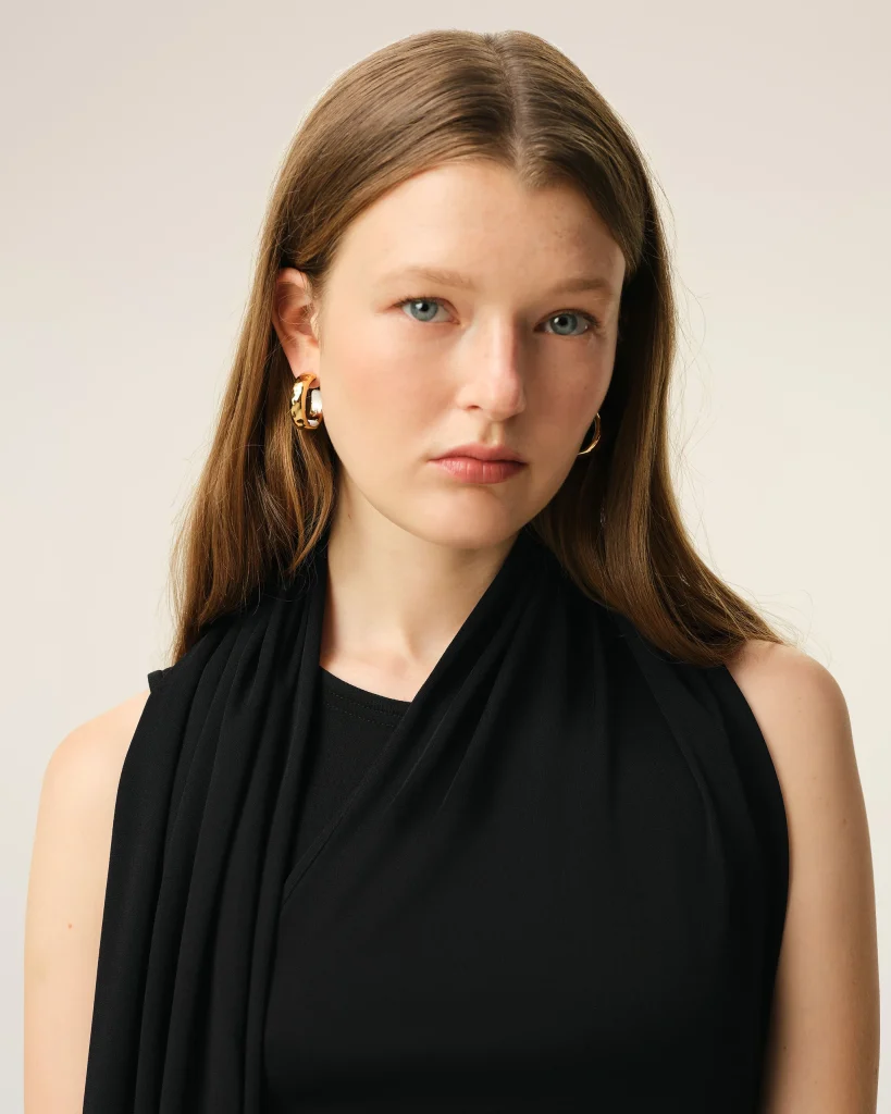 Close-up portrait of a woman with blue eyes, wearing gold hoop earrings and a black draped top against a beige background.