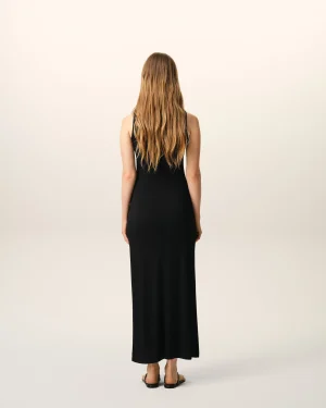 Back view of a woman wearing a black sleeveless dress, with long wavy blonde hair, standing in a neutral studio background.