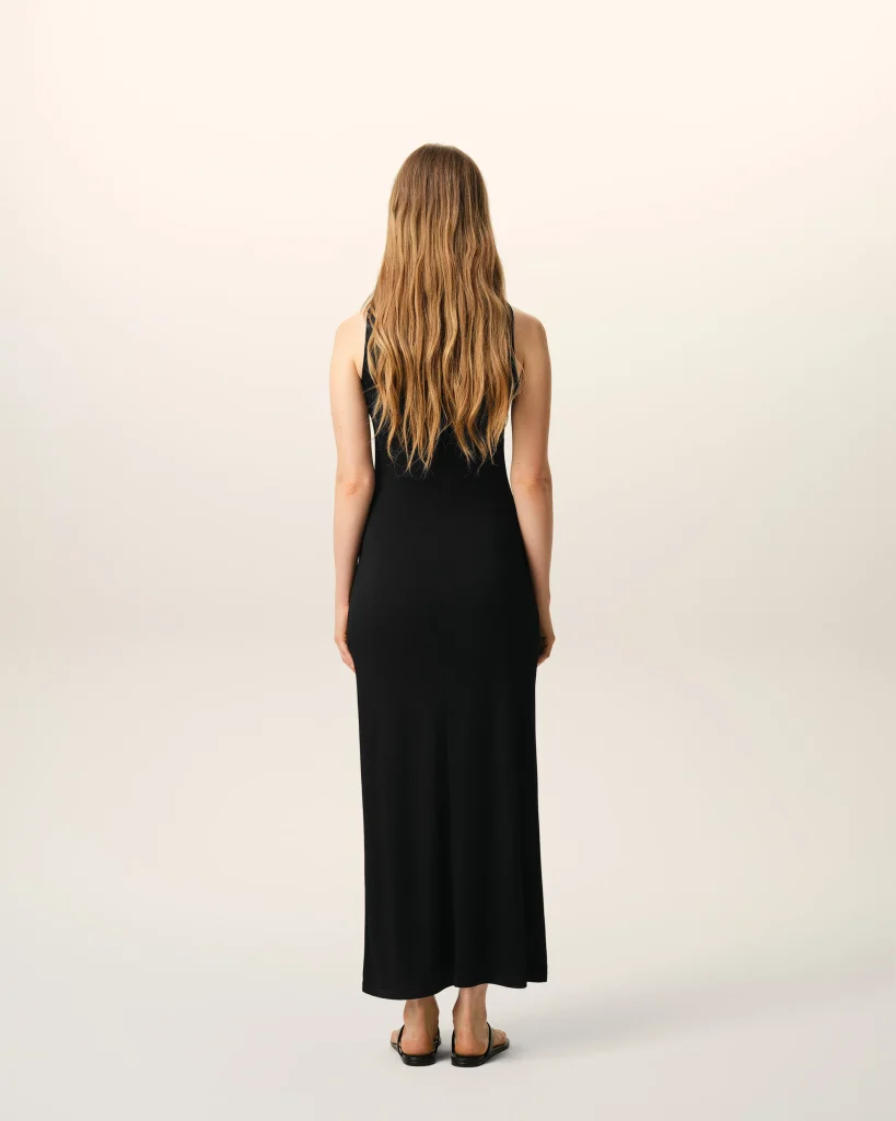 Back view of a woman wearing a black sleeveless dress, with long wavy blonde hair, standing in a neutral studio background.