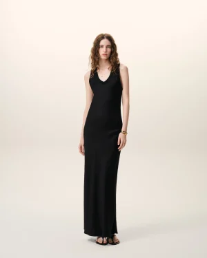 Full-body portrait of a woman with wavy brown hair wearing a black sleeveless maxi dress and sandals, standing against a pale beige backdrop with a calm expression.