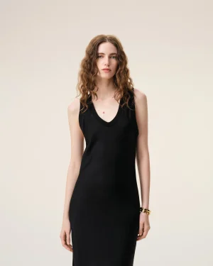 Woman with wavy auburn hair wearing a black sleeveless dress, standing against a light beige background looking at the camera.