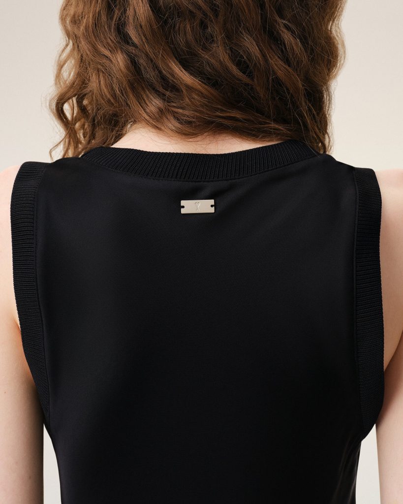 Back view of a person with wavy brown hair wearing a black sleeveless jacket; a small metal tag sits at the upper back center.
