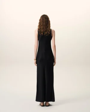 Person seen from behind, wearing a long black sleeveless dress with curly brown hair, standing against a pale gradient background.