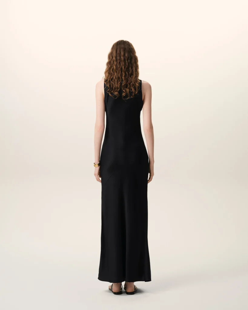 Person seen from behind, wearing a long black sleeveless dress with curly brown hair, standing against a pale gradient background.