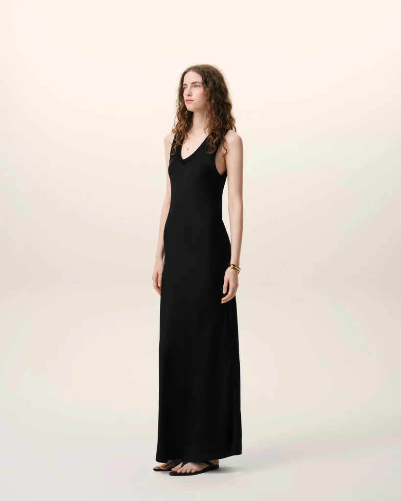 Full-body portrait of a young woman in a black sleeveless maxi dress, standing barefoot against a pale studio backdrop, looking slightly to the left with a neutral expression.