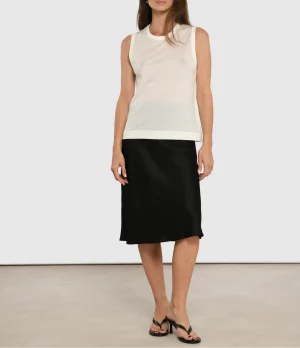 Model wearing a white sleeveless top and a black midi skirt with black sandals, standing against a neutral backdrop.