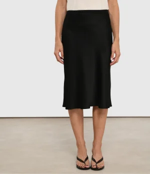 Model wearing a black midi skirt with an elastic waistband, paired with a white top and black strappy sandals, standing on a light floor.