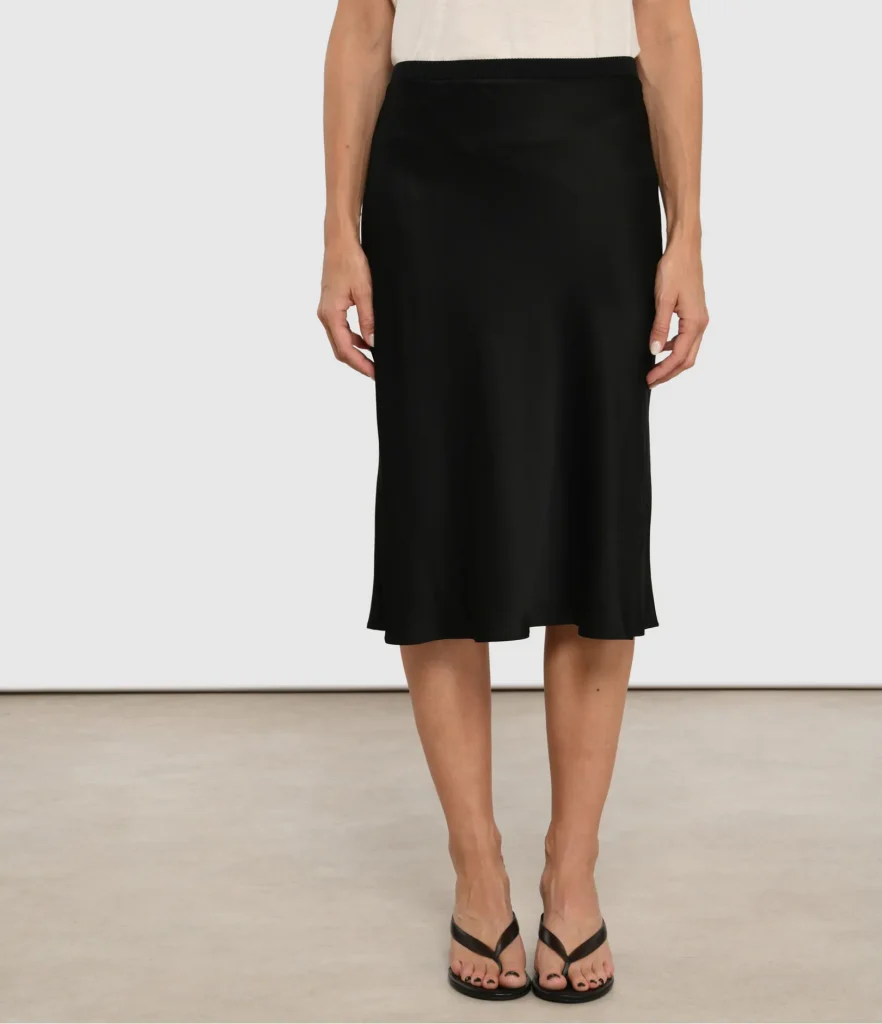Model wearing a black midi skirt with an elastic waistband, paired with a white top and black strappy sandals, standing on a light floor.