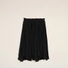 Black pleated skirt hangs against a pale beige wall, displaying its elastic waistband and flowing fabric.