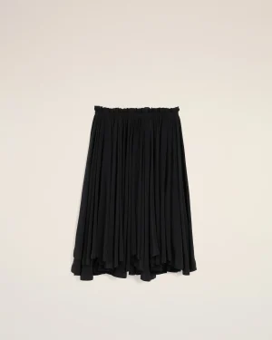 Black pleated skirt hangs against a pale beige wall, displaying its elastic waistband and flowing fabric.