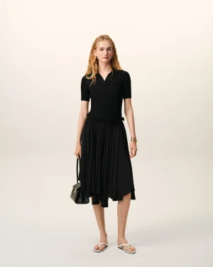 Model standing in a black midi dress with short sleeves, holding a small black handbag against a pale backdrop.