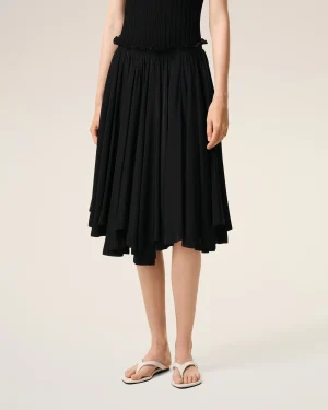 Lower half of a person wearing a black pleated midi skirt with a ruffled waist, standing in light background, wearing beige flip-flops.