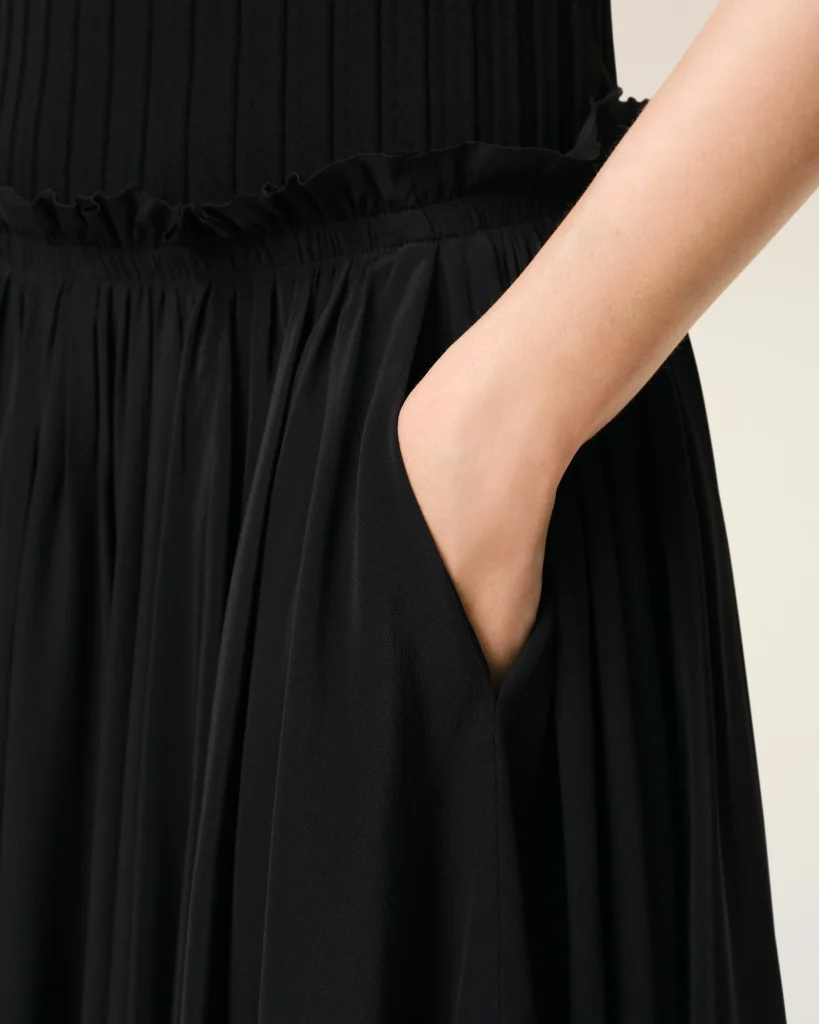 Close-up of a black pleated dress with an elastic waist and an arm resting beside it, beige background