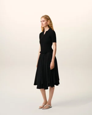 Young woman with blonde hair wearing a black ribbed polo dress and white sandals, standing against a pale gradient background, facing slightly left.