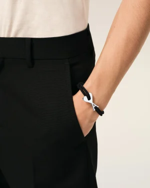 Close-up of a wrist wearing a black cord bracelet with a silver scissor charm, worn against black pants and a white shirt