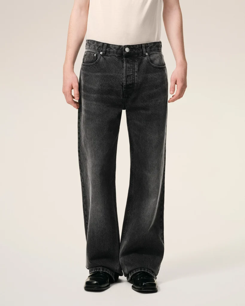 Lower torso and legs of a person wearing dark wash jeans and black shoes against a beige background, front view.