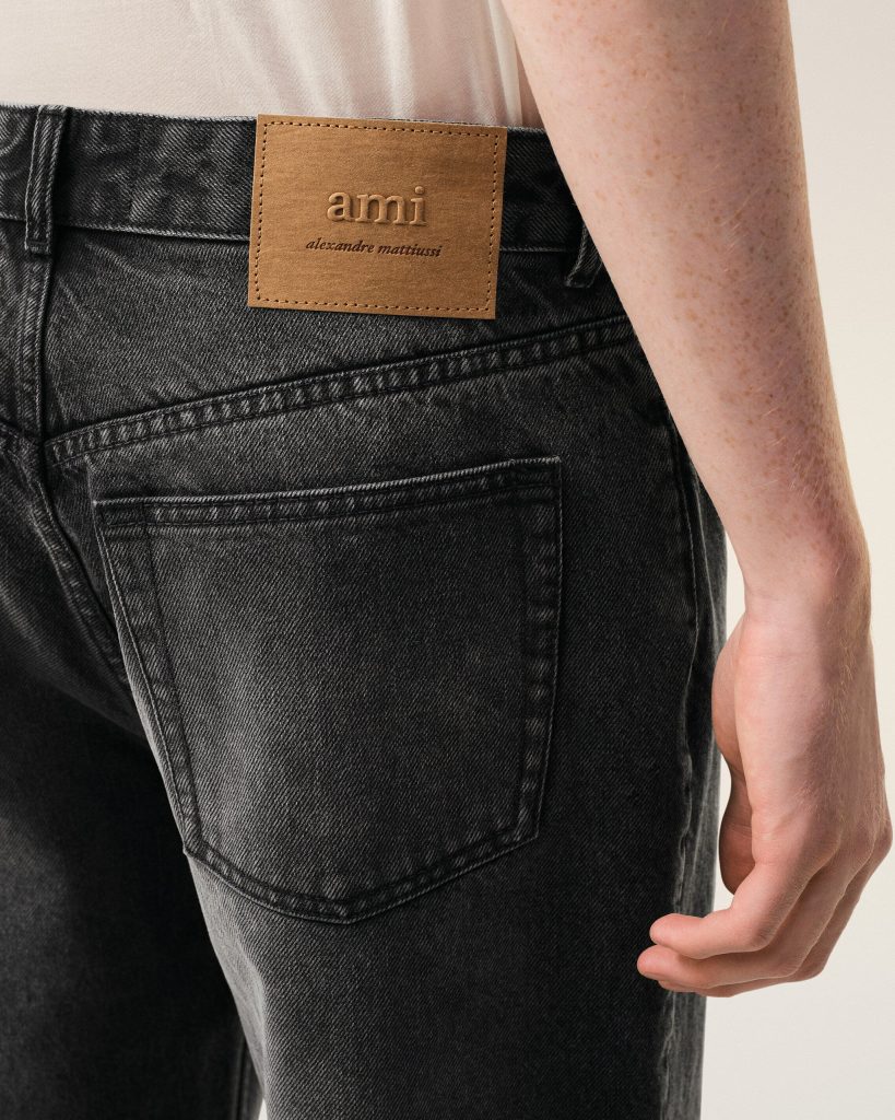 Close-up of a back pocket on dark denim jeans with a tan leather patch reading 'ami' and 'alexandre mattiussi'.