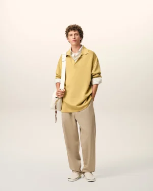 Full-body portrait of a man in a beige oversized top, tan pants, white sneakers, and a white shoulder bag against a pale background