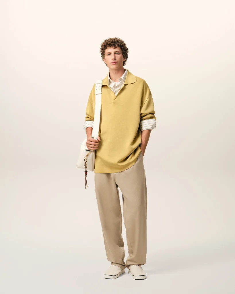 Full-body portrait of a man in a beige oversized top, tan pants, white sneakers, and a white shoulder bag against a pale background