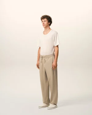 Full-body shot of a young man standing in a neutral studio wearing a white T-shirt, beige drawstring pants, and white sneakers.