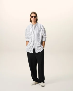 Person standing against a light background, wearing a loose striped button-down shirt, black pants, white sneakers, and dark sunglasses.